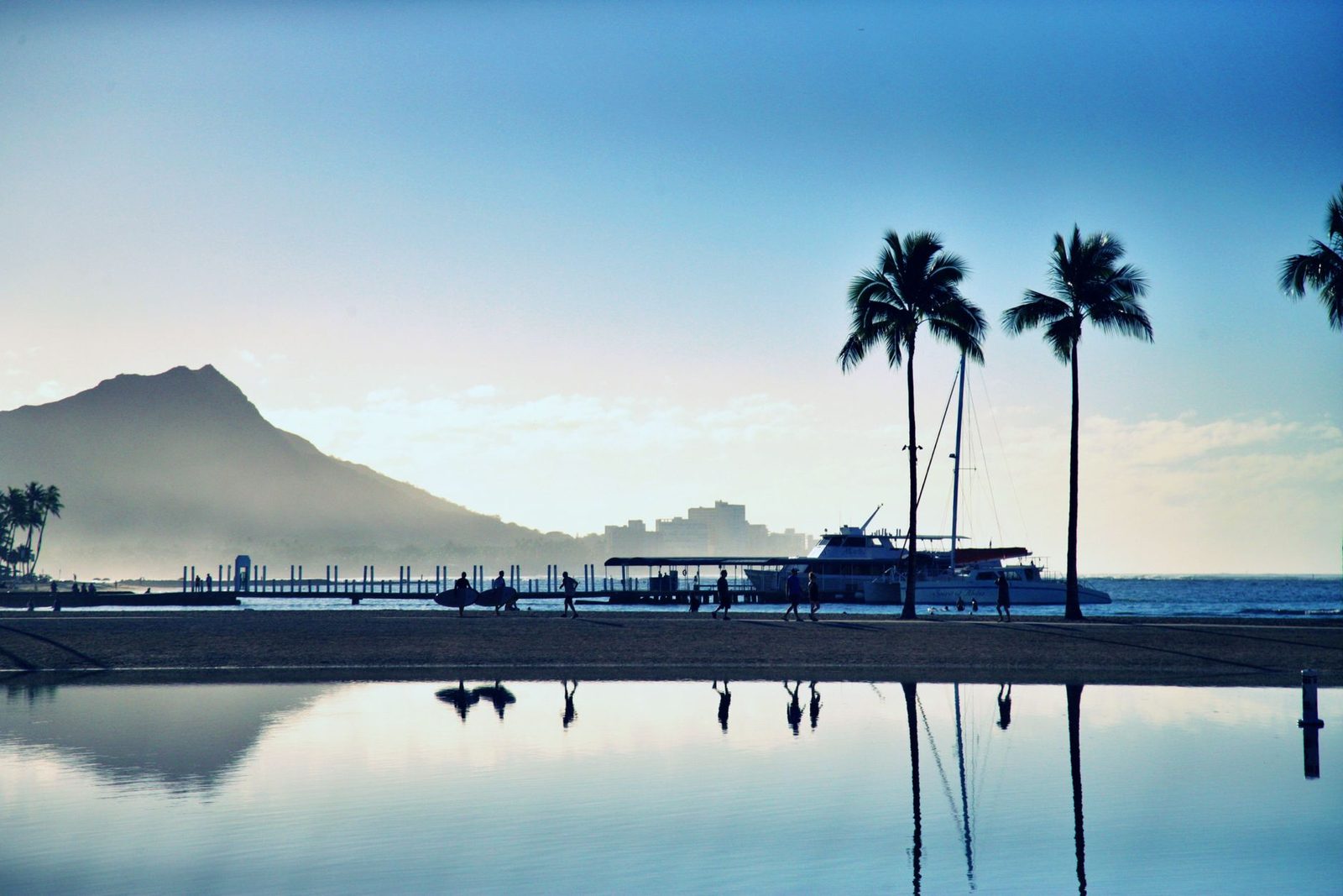 A Hawaiian beach at sunset with palm silhouettes, a moored sailboat and a mountain profile behind